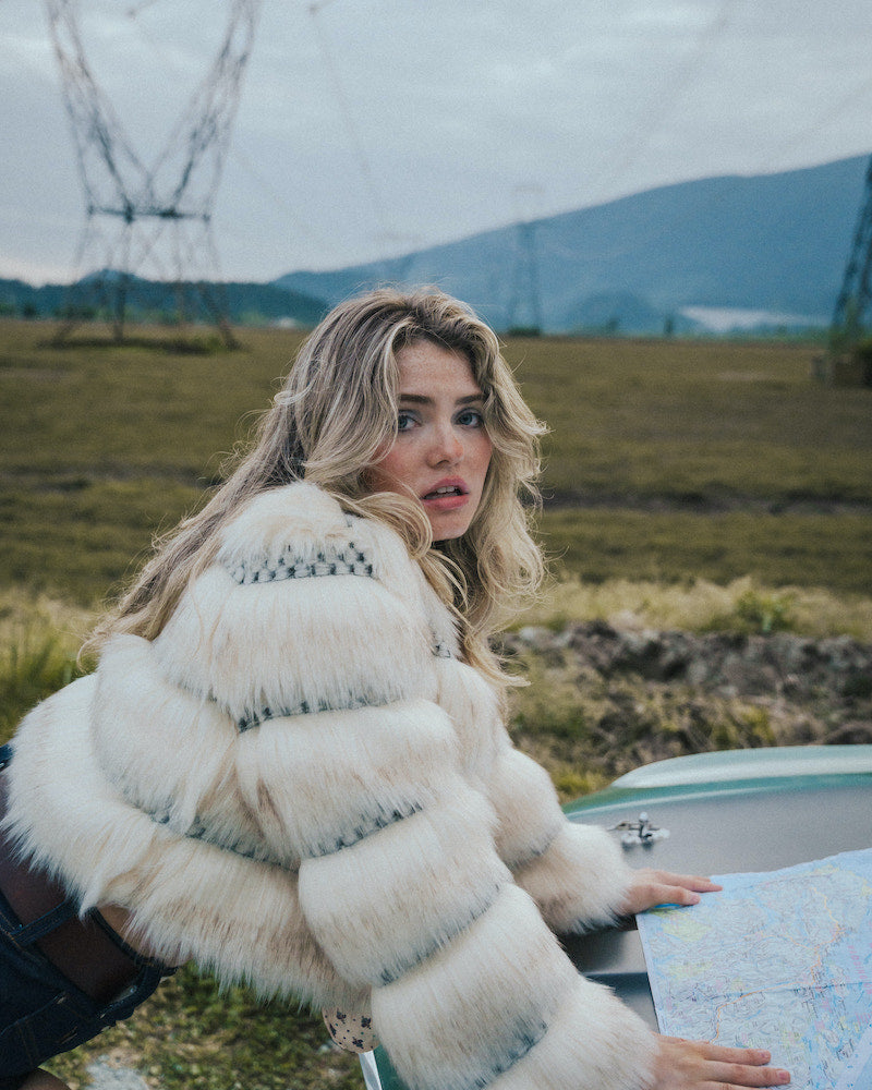 Woman in a white fur coat holding a map outdoors with mountains in the background wearing soul sister since 1969