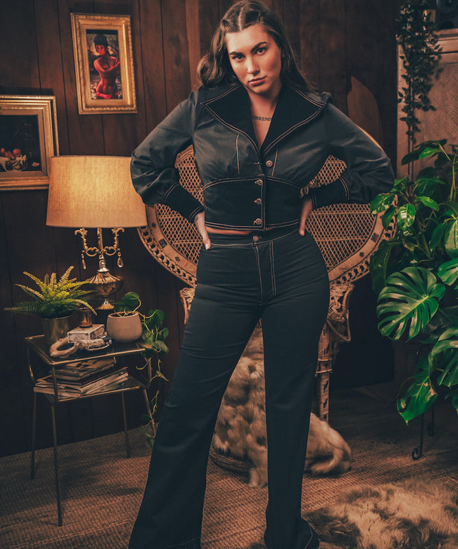 A woman posing in a room with wooden paneling and plants, wearing high rise bell bottom trousers and a cropped jacket.