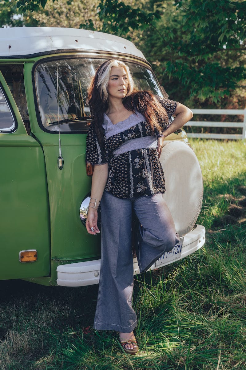 A woman standing beside a green van, wearing a black and white floral print tunic top with long sleeves, paired with gray pants.