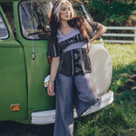 A woman standing beside a green van, wearing a black and white floral print tunic top with long sleeves, paired with gray pants.