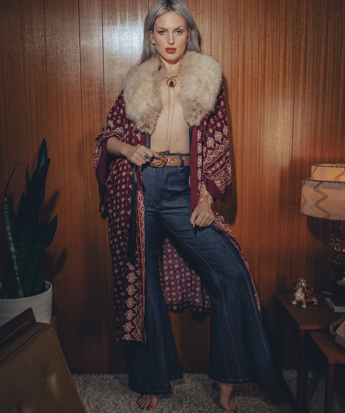 A model wearing high waisted bell bottom jeans and a flowy peasant blouse with a fur stole, standing in a room with a wooden background and a potted plant.