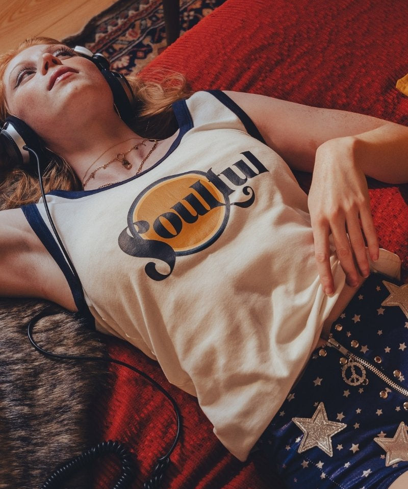 Person wearing headphones  in a 1970s style room and a tank top with a  soulful logo, lying on a red couch.