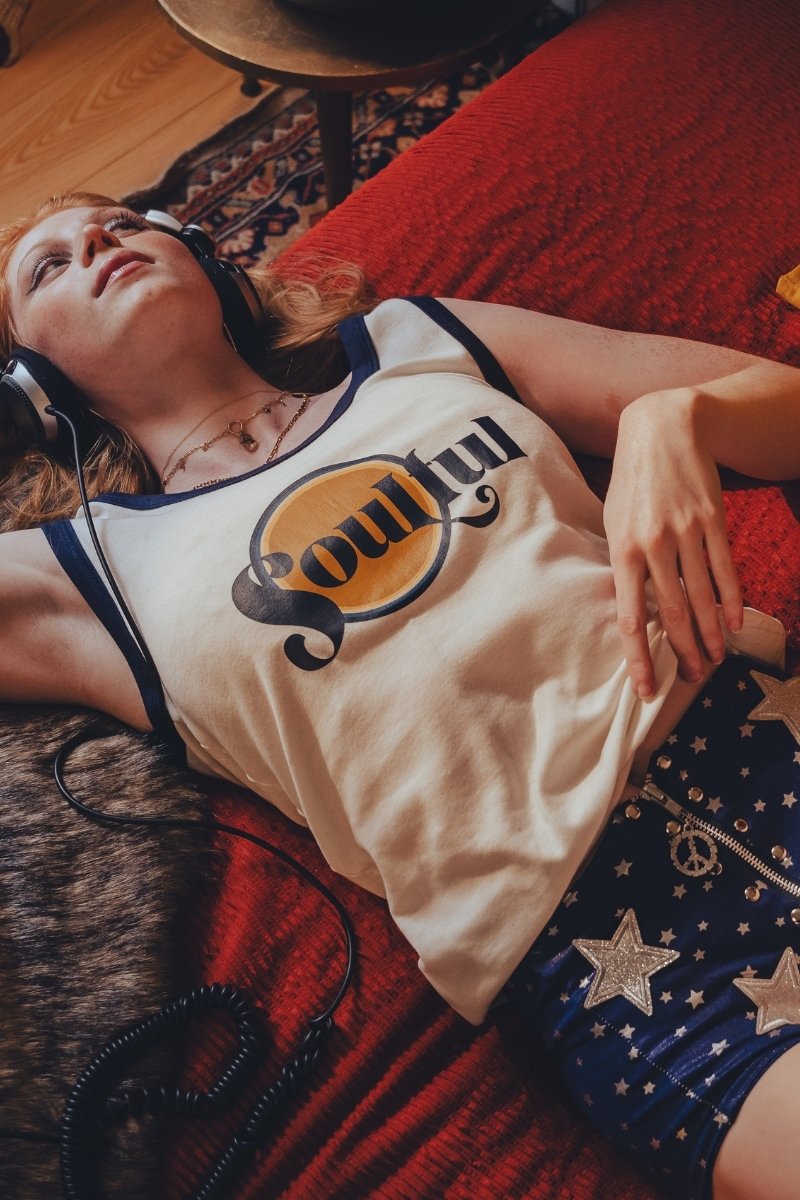 Person wearing headphones  in a 1970s style room and a tank top with a  soulful logo, lying on a red couch.