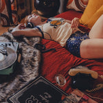 Person lying on a bed wearing a soulful tank top  with a red blanket, in a 1970s styled setting surrounded by books and a plant in a room with patterned curtains.