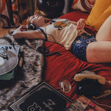 Person lying on a bed wearing a soulful tank top  with a red blanket, in a 1970s styled setting surrounded by books and a plant in a room with patterned curtains.