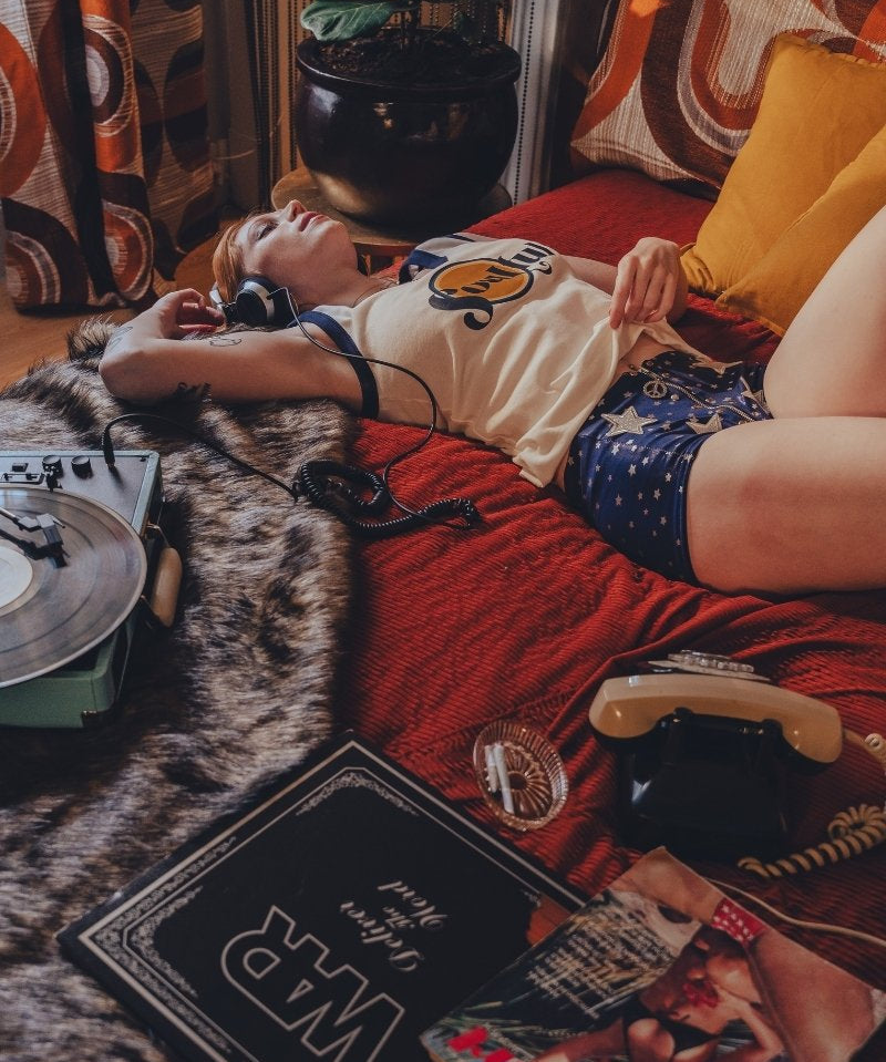 Person lying on a bed wearing a soulful tank top  with a red blanket, in a 1970s styled setting surrounded by books and a plant in a room with patterned curtains.