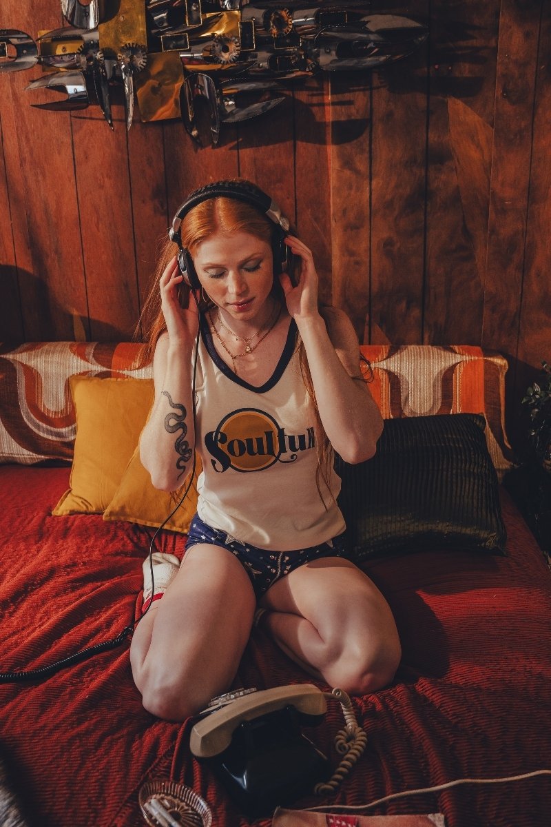 Woman wearing headphones  and a soulful tank top sitting on a couch in a cozy 1970s style room with wooden walls.