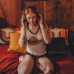 Woman wearing headphones  and a soulful tank top sitting on a couch in a cozy 1970s style room with wooden walls.