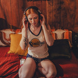 Woman wearing headphones  and a soulful tank top sitting on a couch in a cozy 1970s style room with wooden walls.