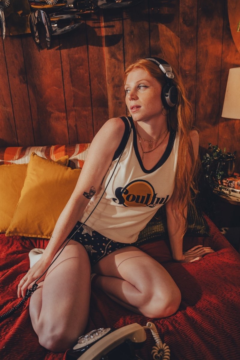 Woman sitting on a bed wearing headphones in a cozy r1970s vibes room with wooden walls.