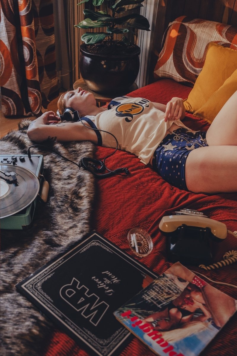 Person lying on a couch with a dog, surrounded by books and a plant in a cozy 1970s style room.