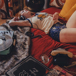 Person lying on a couch with a dog, surrounded by books and a plant in a cozy 1970s style room.