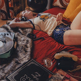 Person lying on a couch with a dog, surrounded by books and a plant in a cozy 1970s style room.