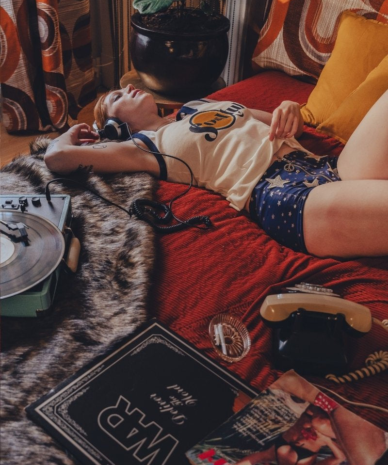 Person lying on a couch with a dog, surrounded by books and a plant in a cozy 1970s style room.