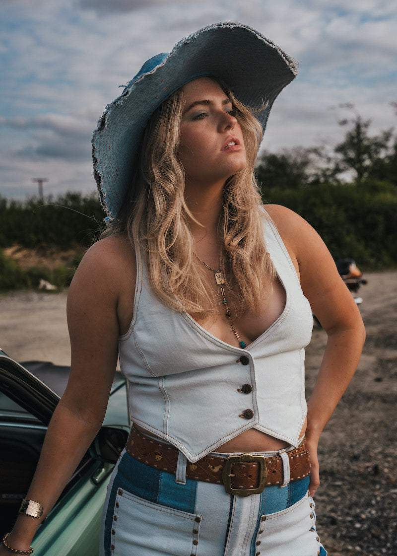 Woman in a white top and blue jeans with a cowboy hat standing next to a car outdoors Wearing Soul Sister since 1969.