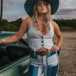 Woman in a white top and blue jeans standing next to a green car on a dirt road Wearing Soul Sister since 1969.