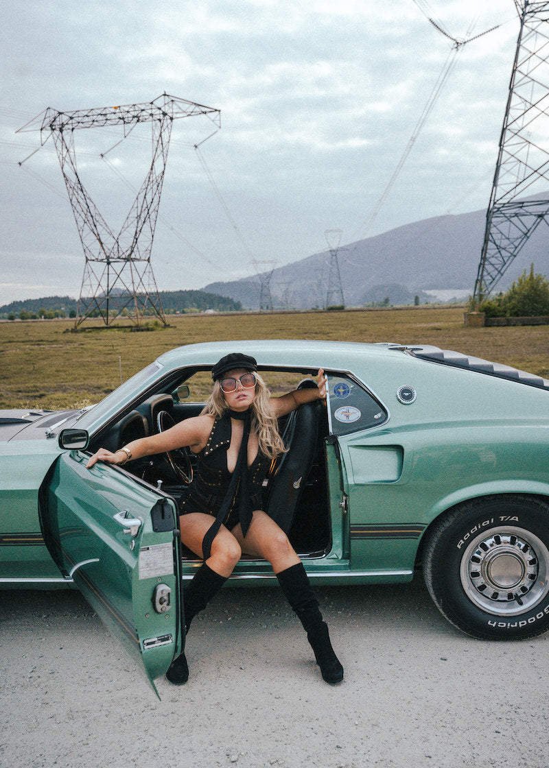 Woman in a black outfit sitting in a green car with power lines and mountains in the background wearing Soul Sister since 1969