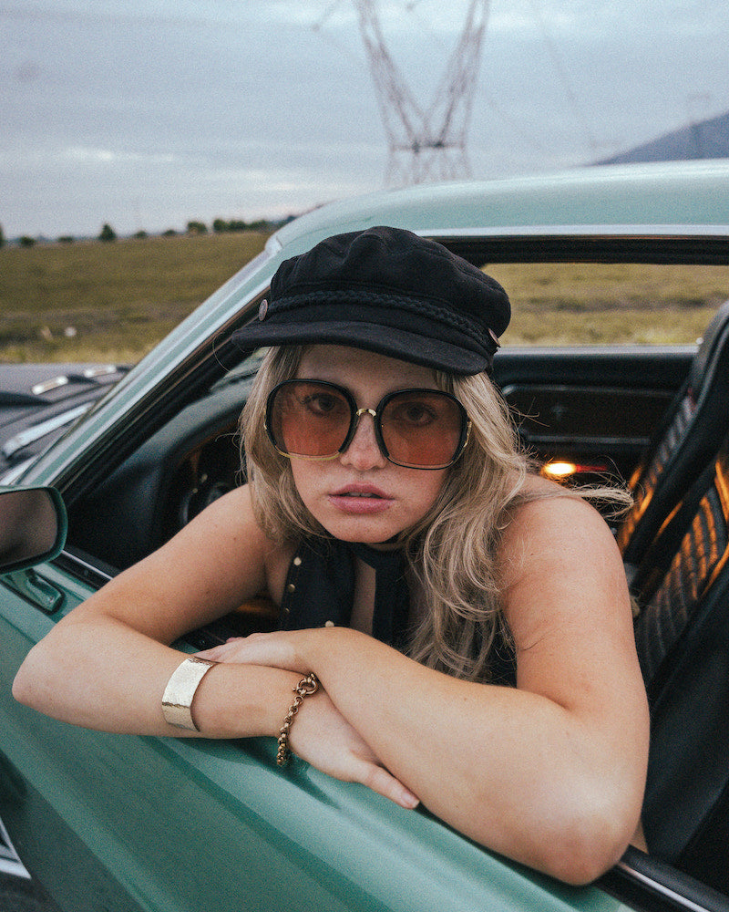 Person leaning out of a car window with sunglasses and a cap, with a scenic background wearing Soul Sister since 1969