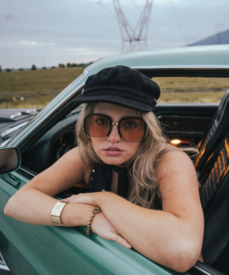 Person leaning out of a car window with sunglasses and a cap, with a scenic background wearing Soul Sister since 1969