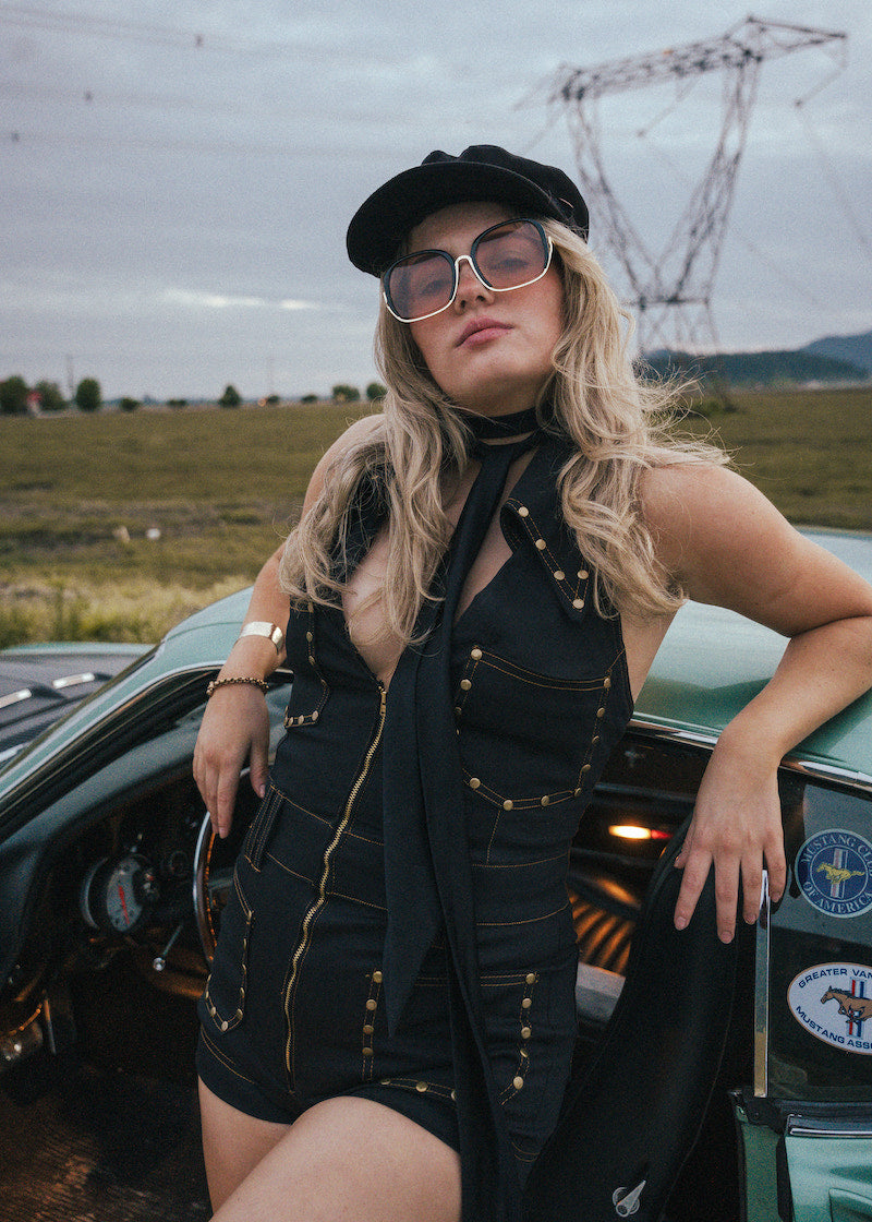 Woman in a black outfit with sunglasses leaning against a vintage car in an open field wearing Soul Sister since 1969.