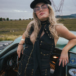 Woman in a black outfit with sunglasses leaning against a vintage car in an open field wearing Soul Sister since 1969.