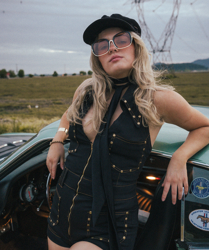 Woman in a black outfit with sunglasses leaning against a vintage car in an open field wearing Soul Sister since 1969.