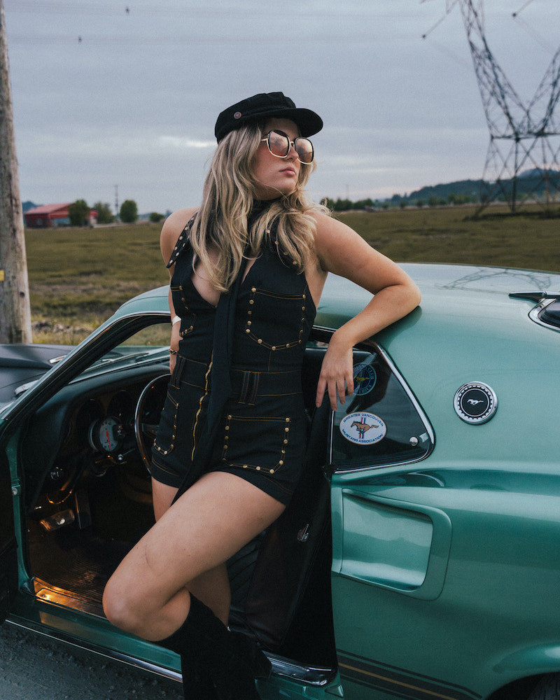 Woman in a black outfit leaning against a vintage green car in an open field wearing Soul Sister since 1969.