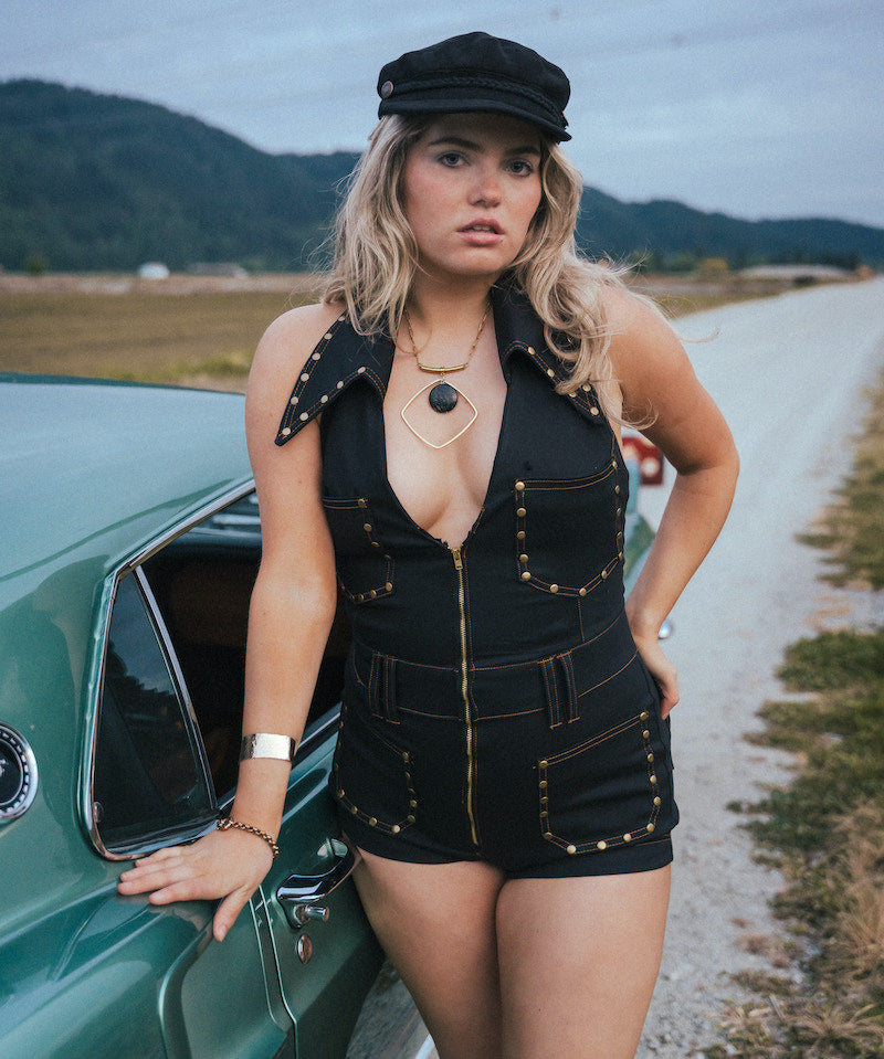 Woman in a black outfit standing next to a vintage car on a rural road wearing Soul Sister since 1969.
