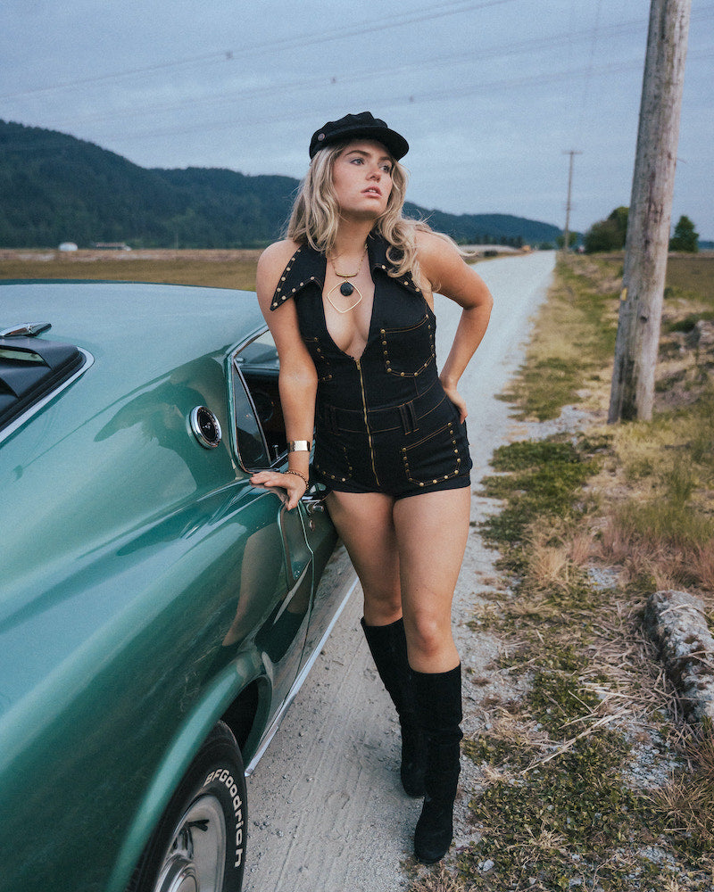 Woman in a black outfit leaning against a green car on a rural road wearing Soul Sister since 1969.