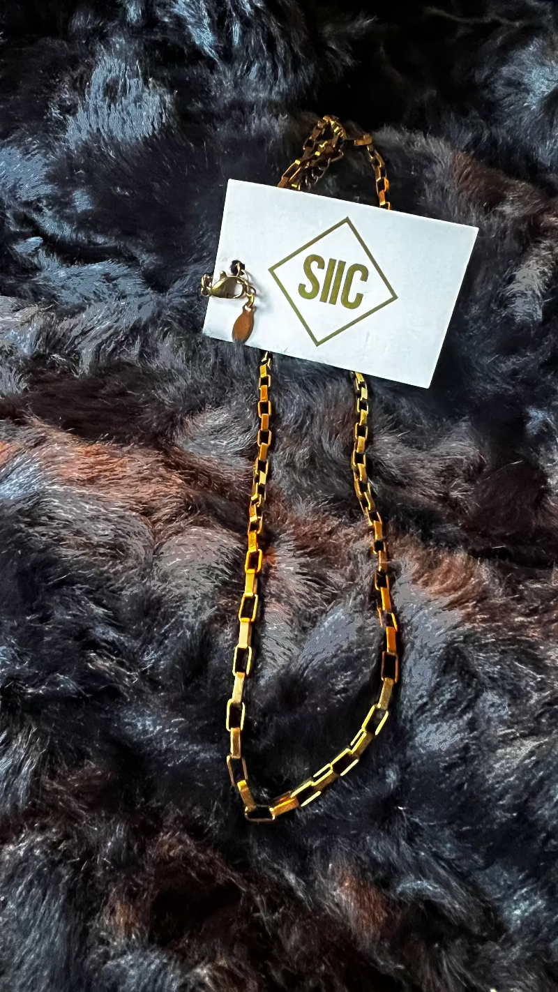 Gold chain necklace on a card with 'SILC' branding, placed on a textured dark fabric background.