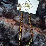 Gold chain necklace on a card with 'SILC' branding, placed on a textured dark fabric background.