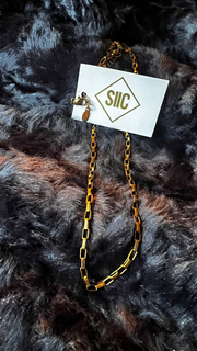 Gold chain necklace on a card with 'SILC' branding, placed on a textured dark fabric background.