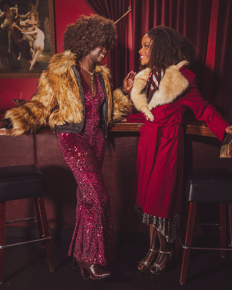 Two women in glamorous  studio 54 disco outfits standing in a bar with red walls and a painting in the background.