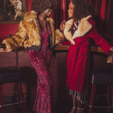 Two women in glamorous  studio 54 disco outfits standing in a bar with red walls and a painting in the background.