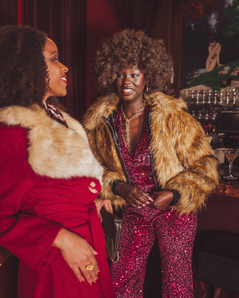 Two women in glamorous  studio 54 70s outfits with fur collars in a bar setting.