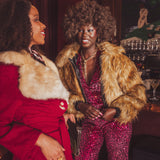 Two women in glamorous  studio 54 70s outfits with fur collars in a bar setting.