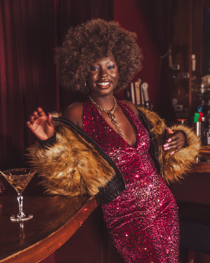 Woman in a red sequin studio 54 style jumpsuit with a  70s inspired fur coat standing at a bar.
