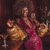 Woman in a red sequin studio 54 style jumpsuit with a  70s inspired fur coat standing at a bar.