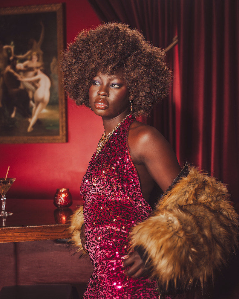 Woman in a red sequin studio 54 style disco jumpsuit with 70s style fur holding a cocktail in a dimly lit room.