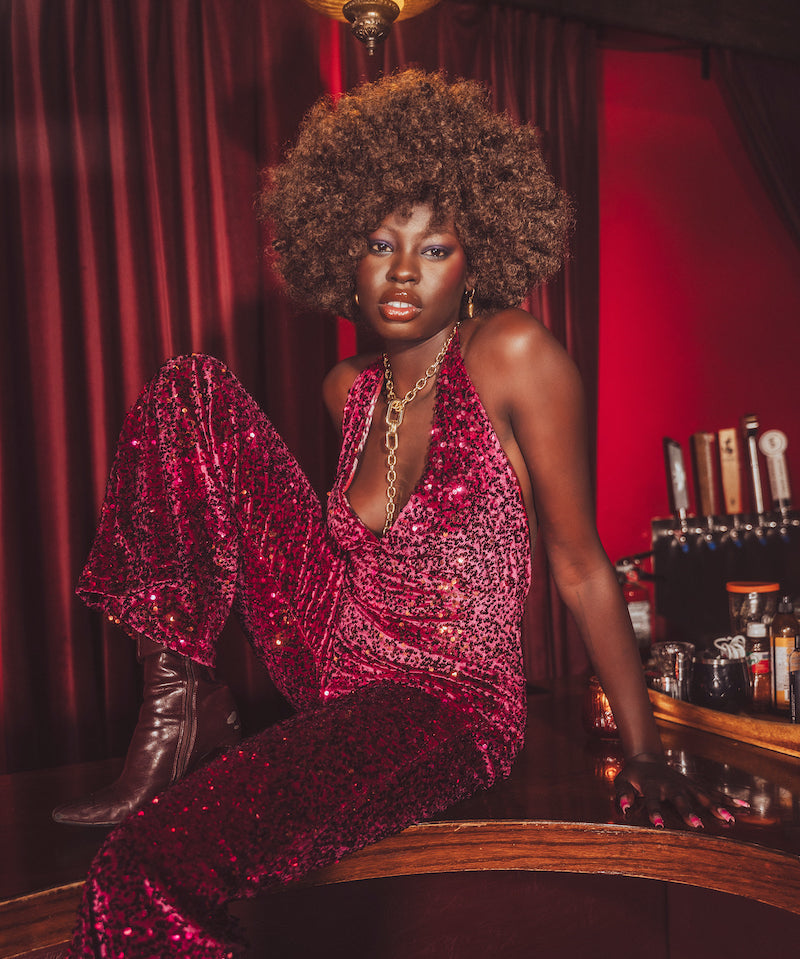 Woman in a red sequin studio 54 style retro jumpsuit  sitting at a bar with red curtains and bottles in the background - 70s inspired clothing made in small batches in Vancouver Canada- Soul Sister since 1969