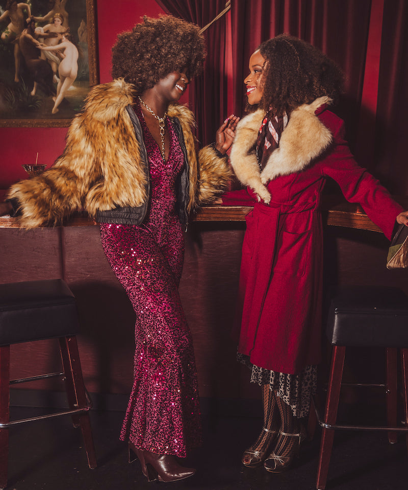 Two women in glamorous outfits - a studio 54 style disco jumpsuit and a fur bombet jacket standing in a bar with red walls and artwork.