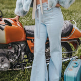 Person wearing a light blue fur coat and jeans standing next to an orange motorcycle with a teal suitcase on grass wearing Soul Sister since 1969.