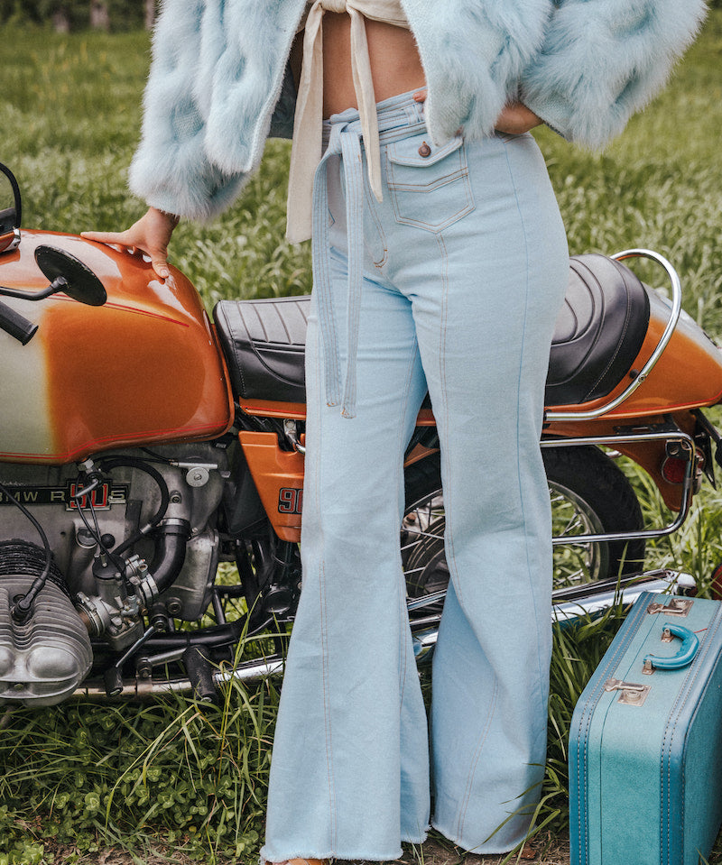 Person wearing a light blue fur coat and jeans standing next to an orange motorcycle with a teal suitcase on grass wearing Soul Sister since 1969.