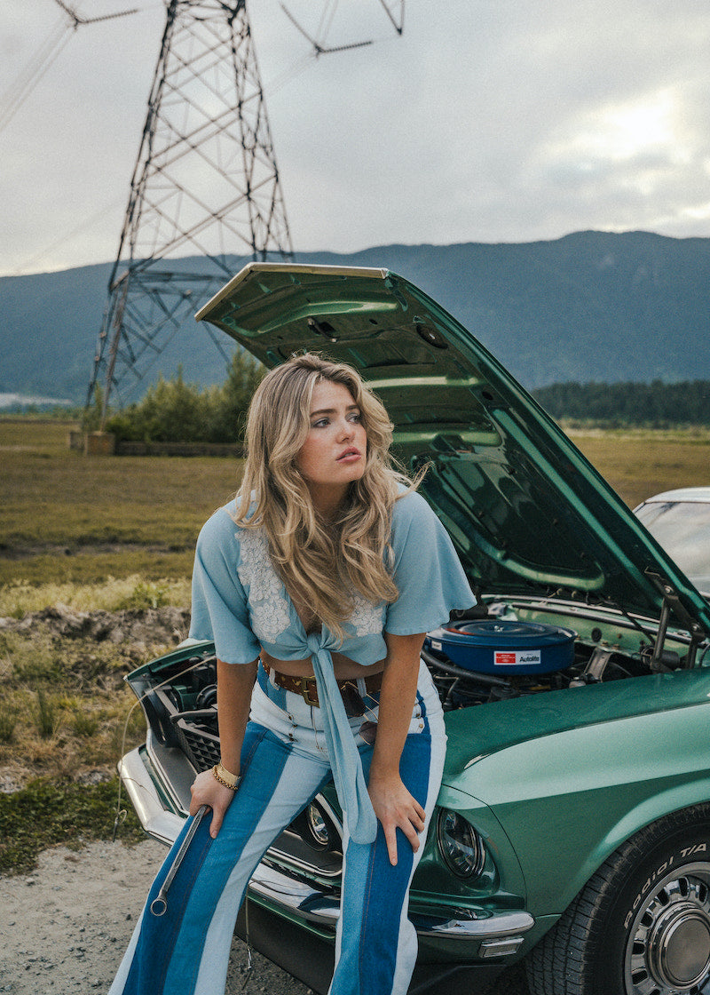 Woman standing next to a vintage car with its hood open in an outdoor setting Wearing Soul Sister since 1969. 
