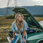 Woman standing next to a vintage car with its hood open in an outdoor setting Wearing Soul Sister since 1969. 