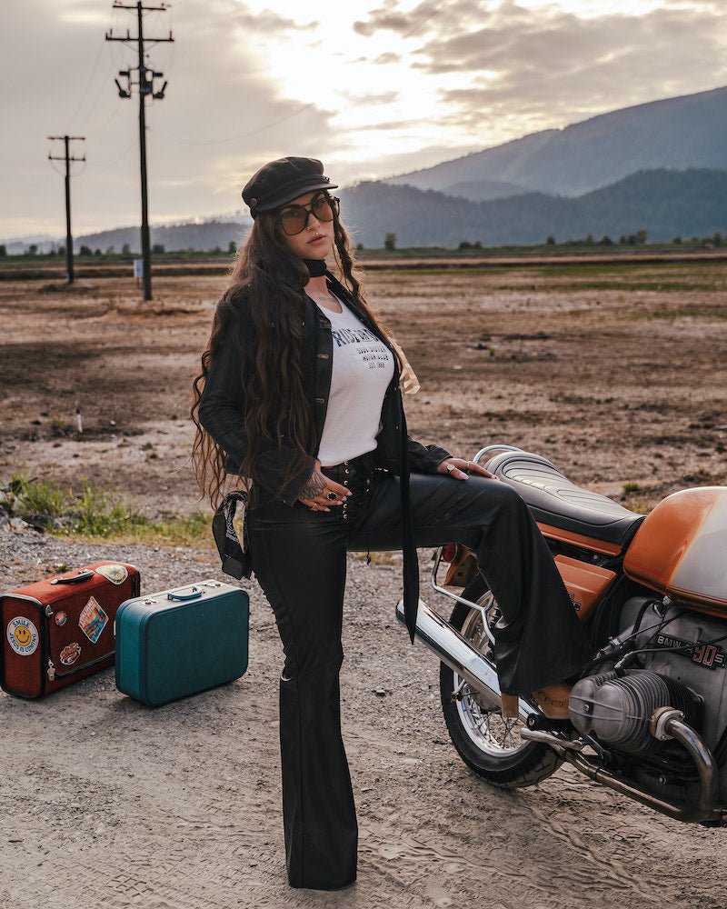 Woman sitting on a motorcycle with suitcases nearby, set against a scenic background wearing Soul Sister since 1969.