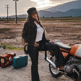 Woman sitting on a motorcycle with suitcases nearby, set against a scenic background wearing clothing designed and made by Soul Sister since 1969..