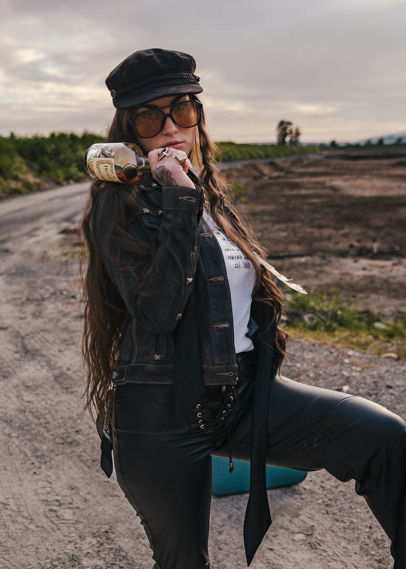Woman in a black jacket and hat holding a bottle in an open field wearing Soul Sister since 1969.