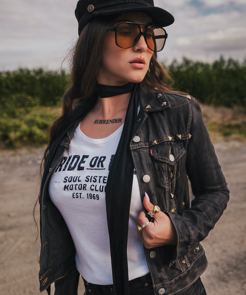Woman wearing a black jacket, white t-shirt, and sunglasses  and a black hat outdoors wearing Soul Sister since 1969.