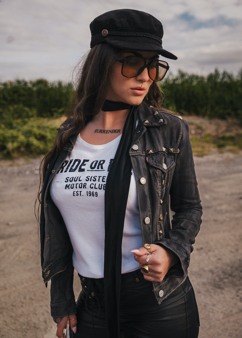 Woman wearing a black jacket, white t-shirt with text, and sunglasses outdoors. wearing Soul Sister since 1969.